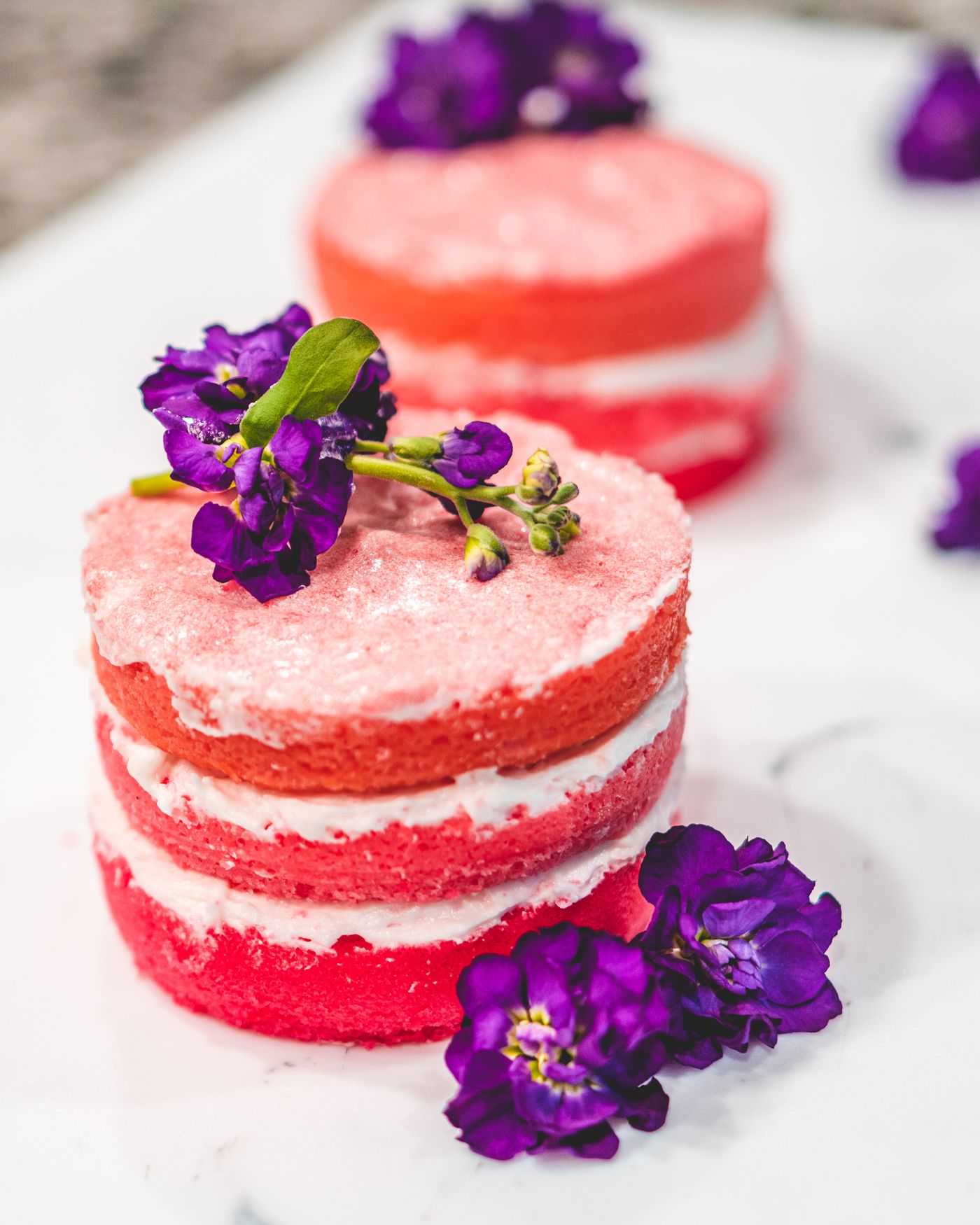 Small frosted layer cakes decorated with buttercream swirls and fresh purple flowers.